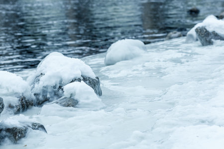 A rocky shoreline with a body of water covered in ice. The rocks are covered in snow, and the water...