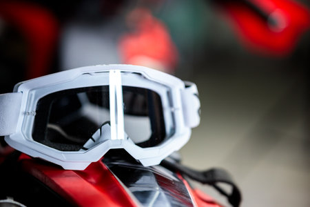 A pair of goggles sits on a red surface. The goggles are white and black with a black strap