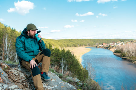 A man sits on a rock by a river, drinking from a water bottle. The scene is peaceful and serene,...