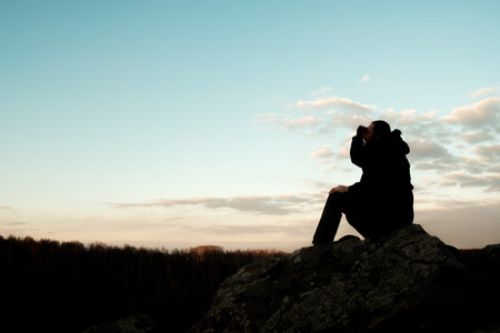 The silhouette of a man sits on a rock, towering over the field. The sky is clear, the sun is setting, creating a peaceful and serene atmosphere.の写真素材