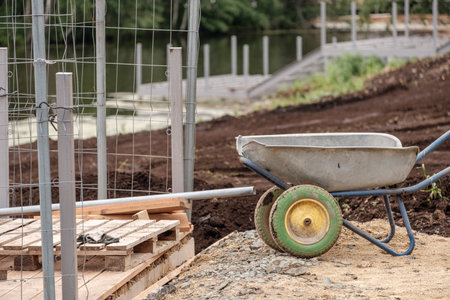 A wheelbarrow is sitting on a dirt field next to a fence. The wheelbarrow is empty and the fence is...