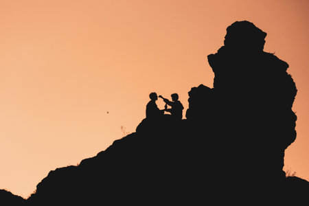 Two people are sitting on a rock. The scene is set against a backdrop of a beautiful sunset,...