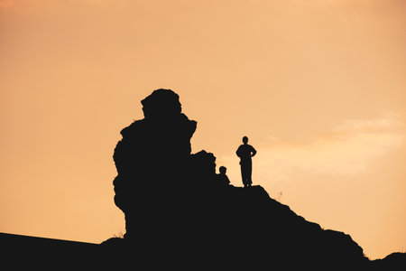A man stands on a rock overlooking a cliff. The sky is orange and the sun is settingの写真素材
