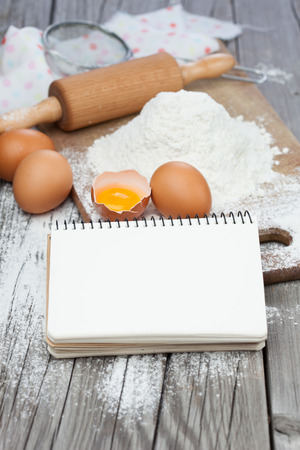 Open recipe book with baking ingredients - flour, eggs and rolling pin on a tableの写真素材