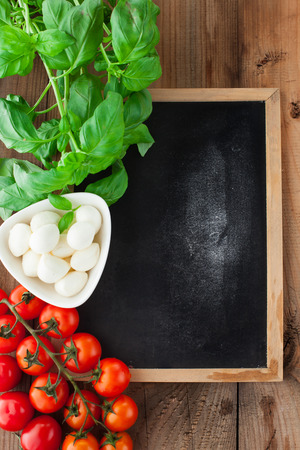 Italian food ingredients - tomatoes, basil, olives, olive oil, garlic, peppercorns and mozzarella on rustic wooden background, with chalkboardの写真素材