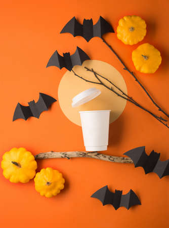 Halloween paper decorations on orange background and cup of coffee to go. Black bats, pumpkins, tree branch and moon. Halloween concept. Flat lay, top view. Party greeting card mockup with copy space.の写真素材