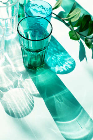 Empty glasses on blue mint paper background. Harsh light through glasses, shadows. Clean minimalist contemporary art. Simple visual concept. AI Aqua trendy color of the year 2021. Selective focus.の写真素材