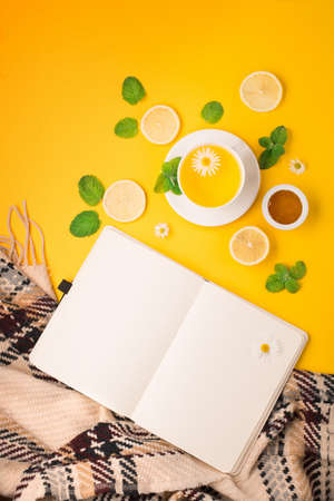 Herbal tea of chamomile flower with mint, lemon, honey and paper notebook on yellow background. Spicy medicinal tea for autumn or winter season. Flat lay, top view. Healthy food and drink concept.の写真素材