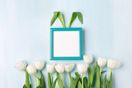 Bunny rabbit ears made of natural green leaves, tulip flowers and empty blue frame on blue wooden background. Easter minimal concept. Flat lay, space for text, modern design template.の写真素材
