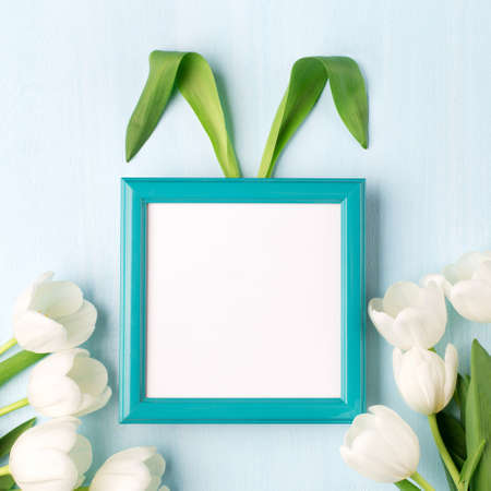 Bunny rabbit ears made of natural green leaves, tulip flowers and empty blue frame on blue wooden...