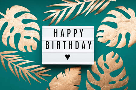 Happy Birthday text on white Lightbox and golden tropical leaves Monstera on paper blue background....