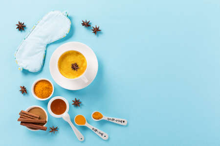 Turmeric golden milk latte with spices, cinnamon, honey and Sleep mask on blue background. Top view, copy space. Moon Milk for better sleep, trendy relaxing bedtime drink. Healthy night sleep concept.の写真素材