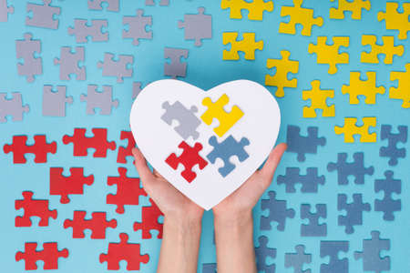 Creative concept design for April 2, Autism World Awareness day. Hands holding large white paper heart and Jigsaw colorful puzzle elements on blue background. Top view, flat lay.の写真素材