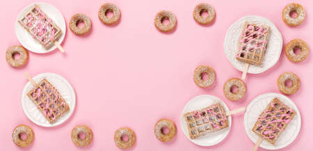 Traditional belgian waffles with sprinkles and delicious donuts with powdered sugar on pastel pink table. Top view, flat lay, copy space. Food background for party, birthday, valentines day.の写真素材