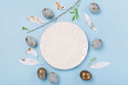 Festive Easter holiday table setting with white empty plate, blue golden eggs and green tree branch on blue background. Happy Easter card concept. Copy space, top view.の写真素材
