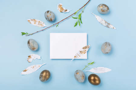 Festive Easter holiday greeting card concept. Easter Decoration with blue golden eggs, empty card and green tree branch on blue background. Copy space, top view.の写真素材