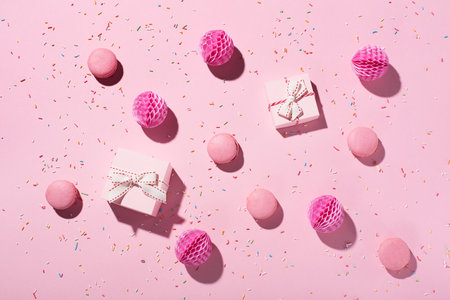 Happy birthday or party background. Flat lay composition with sweet cakes macaroons, sprinkles, pink gift boxes and paper decorations on pink background. Top view, copy space. Festive holiday concept.の写真素材