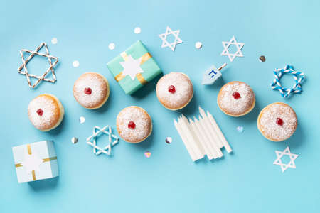 Hanukkah sweet donuts sufganiyot (traditional donuts) with fruit jelly jam, gift boxes, spinnig driedel and candles on blue paper background. Jewish holiday Hanukkah concept. Top view, copy space.の写真素材