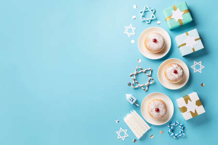 Hanukkah sweet donuts sufganiyot (traditional donuts) with fruit jelly jam, gift boxes, spinnig driedel and candles on blue paper background. Jewish holiday Hanukkah concept. Top view, copy space.の写真素材