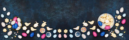 Festive Easter holiday greeting card background. Cute homemade Easter cookies, chocolate eggs, sugar sprinkling on dark blue wooden background. Happy Easter concept. Copy space, top view.の写真素材