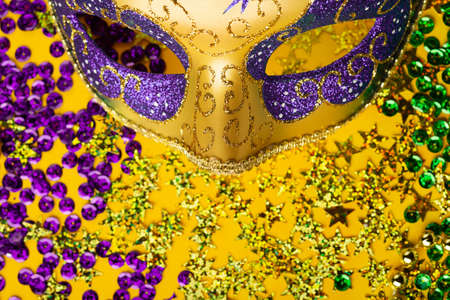 Mardi Gras gold color beads with Luxury Masquerade venitian festival carnival mask and golden, green, purple confetti on yellow background. Party invitation, greeting card, venetian carnivale celebration concept.の写真素材