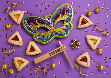 Purim celebration jewish carnival holiday concept. Tasty hamantaschen cookies, Triangular pastry, Festive carnival mask, noisemaker, sweet candies and party decor on purple Background. Top view, flat lay, copy space.の写真素材