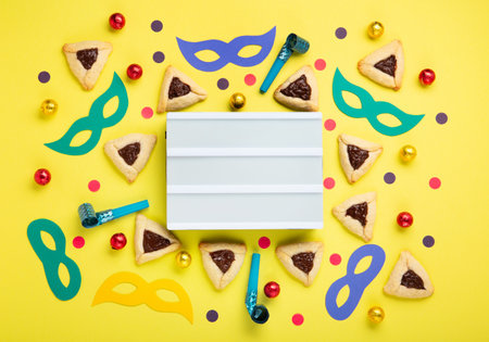 Homemade Purim hamantaschen cookies, Triangular pastry, Carnival mask, noisemaker, sweet candies and festive party decor on yellow background, Top view. Purim celebration jewish holiday concept.の写真素材