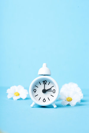 White alarm clock and Daisy Flowers on blue background. Spring forward, Time Change, Daylight Saving Time Ends, Changing the time on the watch to spring time, Summer back concept.の写真素材
