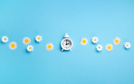 White alarm clock and Daisy Flowers on blue background. Spring forward, Time Change, Daylight Saving Time Ends, Changing the time on the watch to spring time, Summer back concept.の写真素材