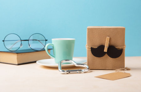 Fathers day holiday card concept with craft gift box, black mustache, cup of coffee, book and glasses on blue background, copy space.の写真素材