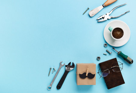 Fathers day holiday card concept with craft gift box, black mustache, cup of coffee, tools, book and glasses on blue background, top view, copy space.の写真素材