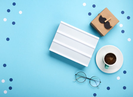 Fathers day holiday card concept with craft gift box, black mustache, cup of coffee, empty white lightbox and glasses on blue background, top view, copy space.の写真素材