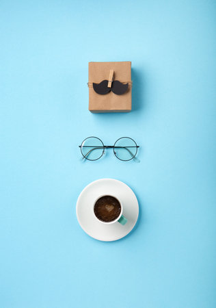 Fathers day holiday card concept with craft gift box, black mustache, cup of coffee and glasses on blue background, top view, copy space.の写真素材