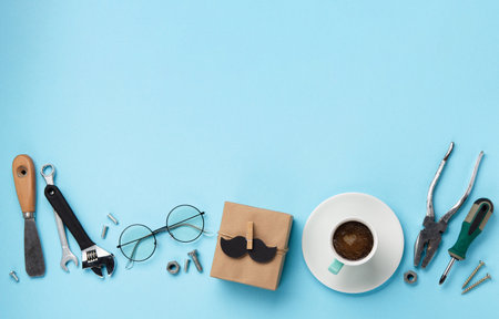 Fathers day holiday card concept with craft gift box, black mustache, cup of coffee, tools and glasses on blue background, top view, copy space.の写真素材