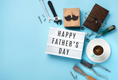 Fathers day holiday card concept with craft gift box, black mustache, cup of coffee, tools, book, empty white lightbox and glasses on blue background, top view, copy space.の写真素材