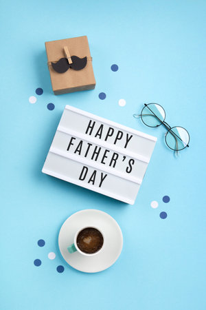 Fathers day holiday card concept with craft gift box, black mustache, cup of coffee, empty white lightbox and glasses on blue background, top view, copy space.の写真素材
