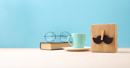 Fathers day holiday card concept with craft gift box, black mustache, cup of coffee, book and glasses on blue background, copy space.の写真素材