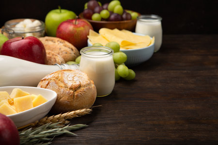 Happy Shavuot holiday card. Jewish religious holiday concept. Dairy products, fruits, cheese, bread, milk bottle, cottage cheese, wheat on dark wooden background, copy space.の写真素材