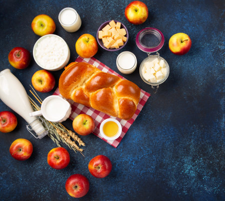 Happy Shavuot holiday card. Jewish religious holiday concept. Dairy products, fresh apples, cheese, bread, milk, cottage cheese, wheat and honey on dark blue wooden background. Top view, copy space.の写真素材