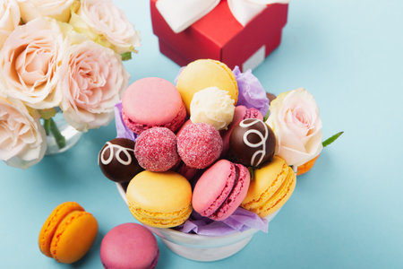 Sweet cookies macaroons, red gift box, rose flowers and chocolate candies on blue background. Spring presents concept for Mother's day, woman's day, birthday. Flat lay, empty space for text or messageの写真素材