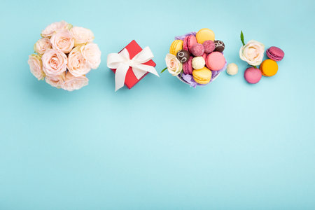 Sweet cookies macaroons, red gift box, rose flowers and chocolate candies on blue background. Spring presents concept for Mother's day, woman's day, birthday. Flat lay, empty space for text or messageの写真素材