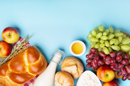 Happy Shavuot holiday card. Jewish religious holiday concept. Dairy products, fresh apples, grapes, cheese, bread, milk, cottage cheese, wheat and honey on blue background. Top view, copy space.の写真素材