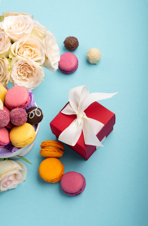 Sweet cookies macaroons, red gift box, rose flowers and chocolate candies on blue background. Spring presents concept for Mother's day, woman's day, birthday. Flat lay, empty space for text or messageの写真素材