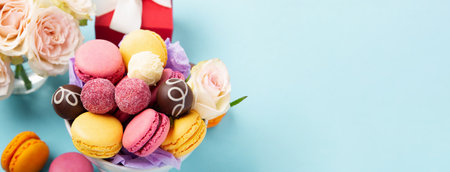 Sweet cookies macaroons, red gift box, rose flowers and chocolate candies on blue background. Spring presents concept for Mother's day, woman's day, birthday. Banner, empty space for text.の写真素材