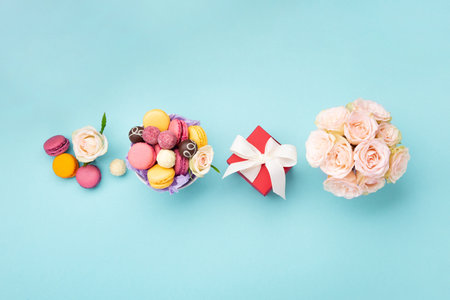 Sweet cookies macaroons, red gift box, rose flowers and chocolate candies on blue background. Spring presents concept for Mother's day, woman's day, birthday. Top view, empty space for textの写真素材