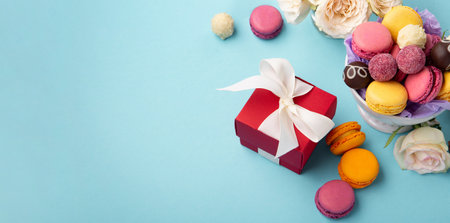 Sweet cookies macaroons, red gift box, rose flowers and chocolate candies on blue background. Spring presents concept for Mother's day, woman's day, birthday. Banner, empty space for textの写真素材