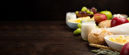Happy Shavuot holiday card. Jewish religious holiday concept. Dairy products, fruits, cheese, bread, milk bottle, cottage cheese, wheat on dark wooden background. Banner, copy space.の写真素材