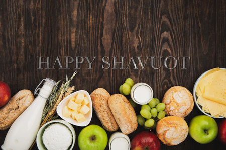 Happy Shavuot holiday card. Jewish religious holiday concept. Dairy products, fruits, cheese, bread, milk bottle, cottage cheese, wheat on dark wooden background. Top view, copy space.の写真素材