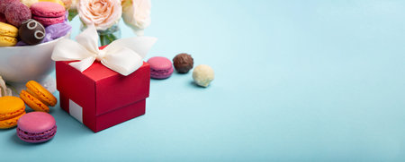 Sweet cookies macaroons, red gift box, rose flowers and chocolate candies on blue background. Spring presents concept for Mothers day, womans day, birthday, empty space for text or messageの写真素材