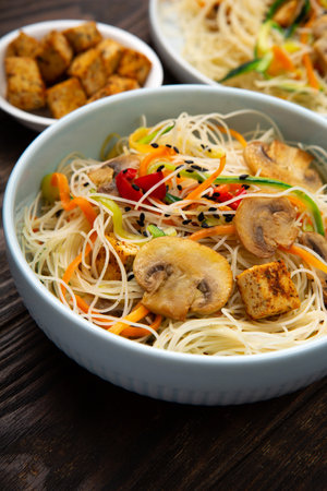 Vegan rice noodles with tofu, peppers, carrots and zucchini prepared for wook on dark brown background. Healthy vegan food conceptの写真素材